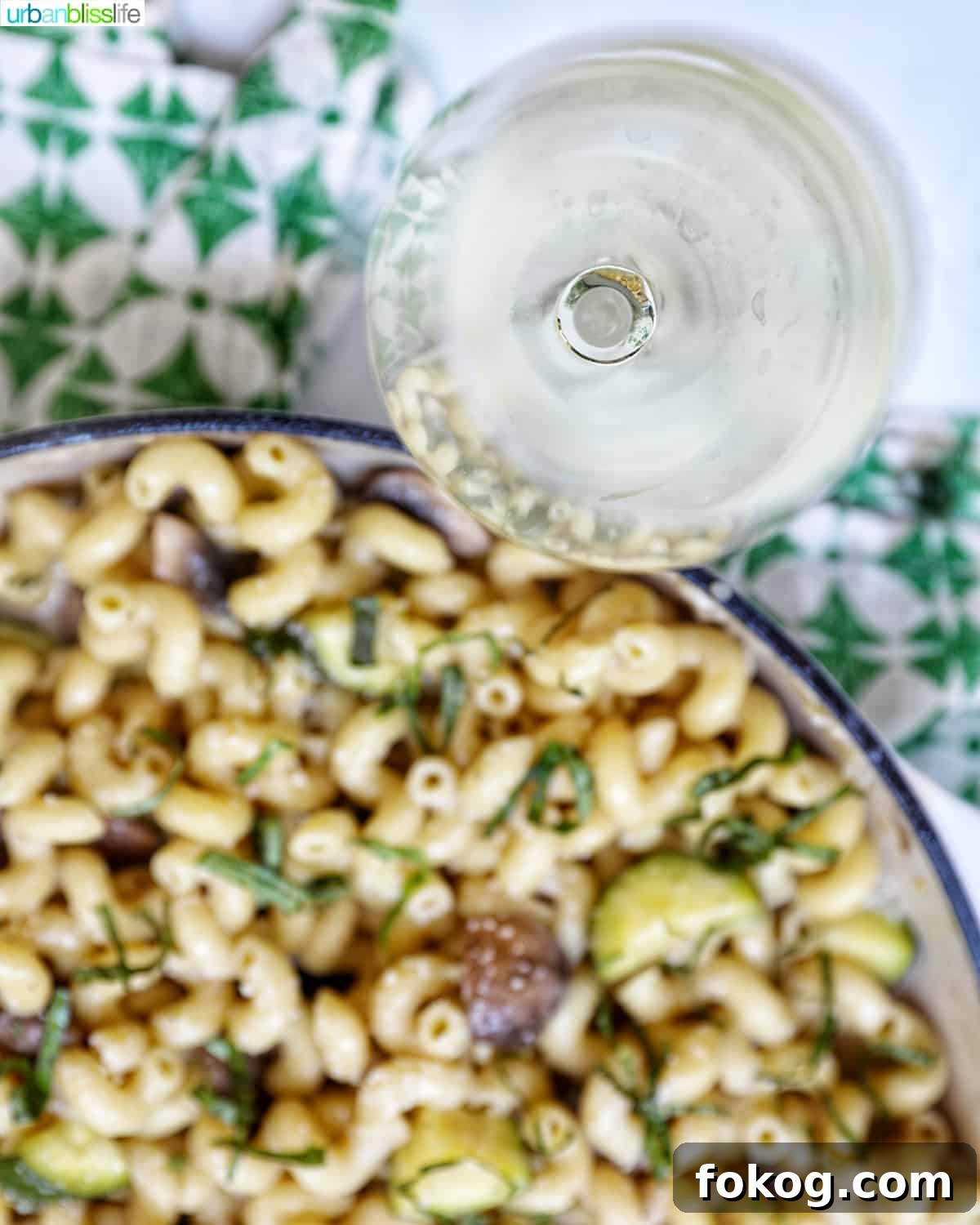 overhead photo of a glass of white wine next to pan of pasta with a green and white napkin.