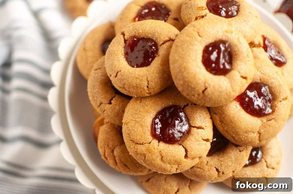 A plate stacked with Peanut butter jelly thumbprint cookies filled with red jam, placed on a white stand next to a striped cloth.