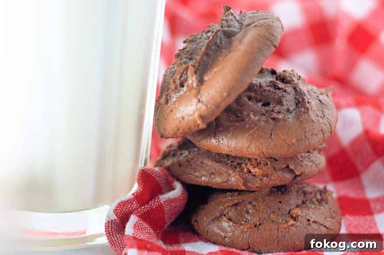 Three Fudgy Chocolate Brownie Cookies stacked beside a glass of milk on a red and white checkered cloth.