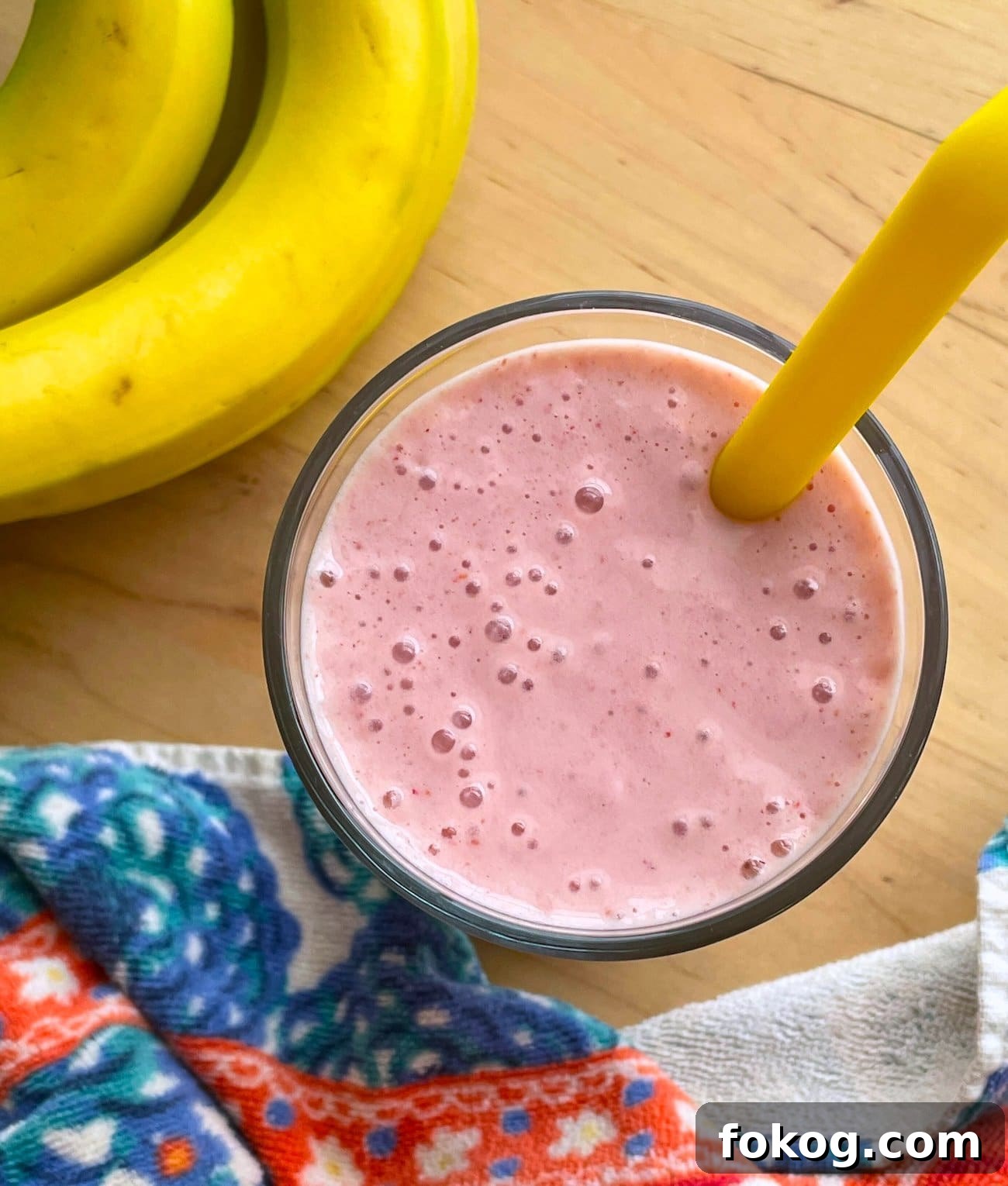 Strawberry Banana Yogurt Smoothie Recipe — Creamy & Refreshing 2 A close-up of a strawberry banana smoothie with a yellow straw with bubbles on top, a flowery towel off to the side, and a pile of bananas.