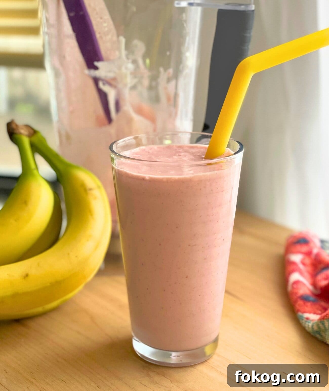 Strawberry Banana Yogurt Smoothie Recipe — Creamy & Refreshing 3 A clear glass of strawberry banana smoothie with a yellow straw, a pile of bananas, and a blender.