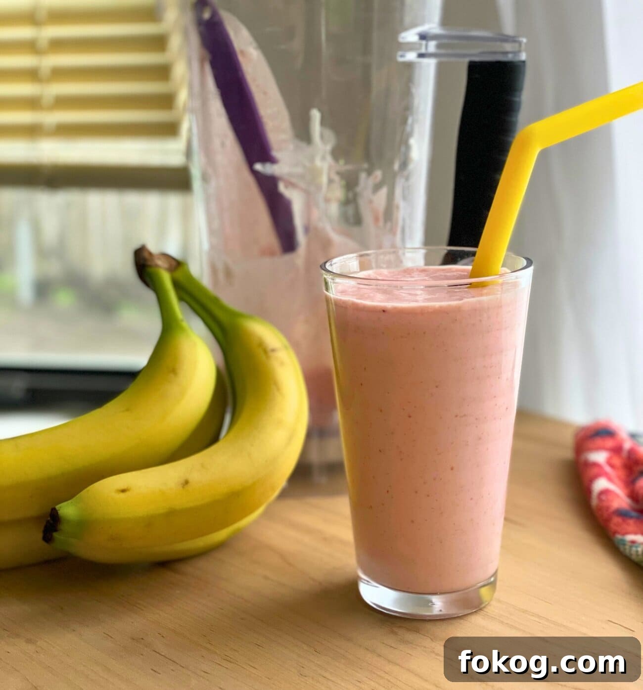 Strawberry Banana Yogurt Smoothie Recipe — Creamy & Refreshing 7 A clear glass of strawberry banana smoothie with a yellow straw, a pile of bananas, and a blender.