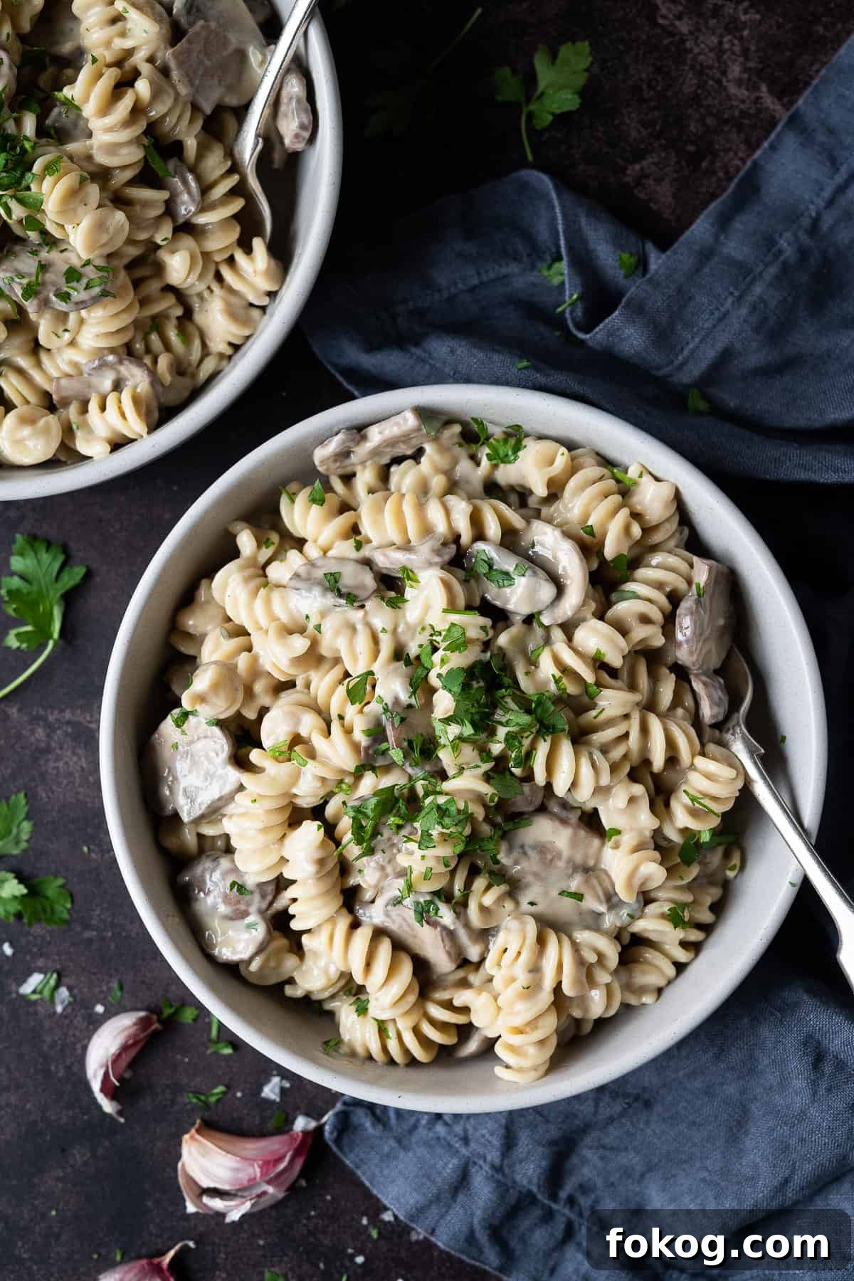 Creamy vegan mushroom pasta with truffle oil - this incredible vegan pasta is rich, creamy and delicious. With a cashew cream sauce and truffle oil it is an indulgent, comforting and impressive meal that is easy to make but tastes amazing! #veganmeal #dairyfree #plantbased Two grey bowls of mushroom pasta on a dark background with a grey cloth, fresh parsley and garlic skins.