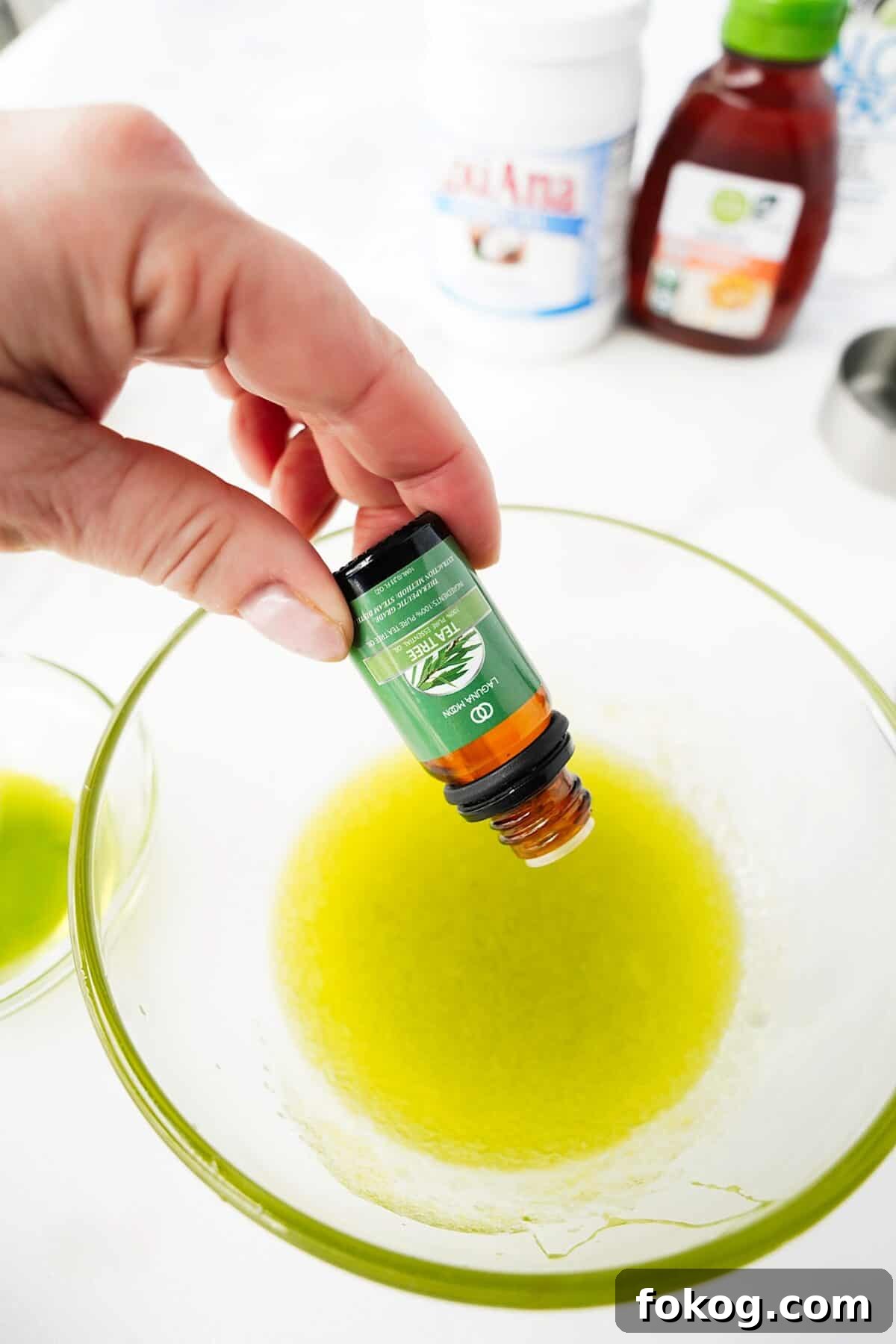 hand adding tea tree oil to glass bowl with strained mashed avocados and oils