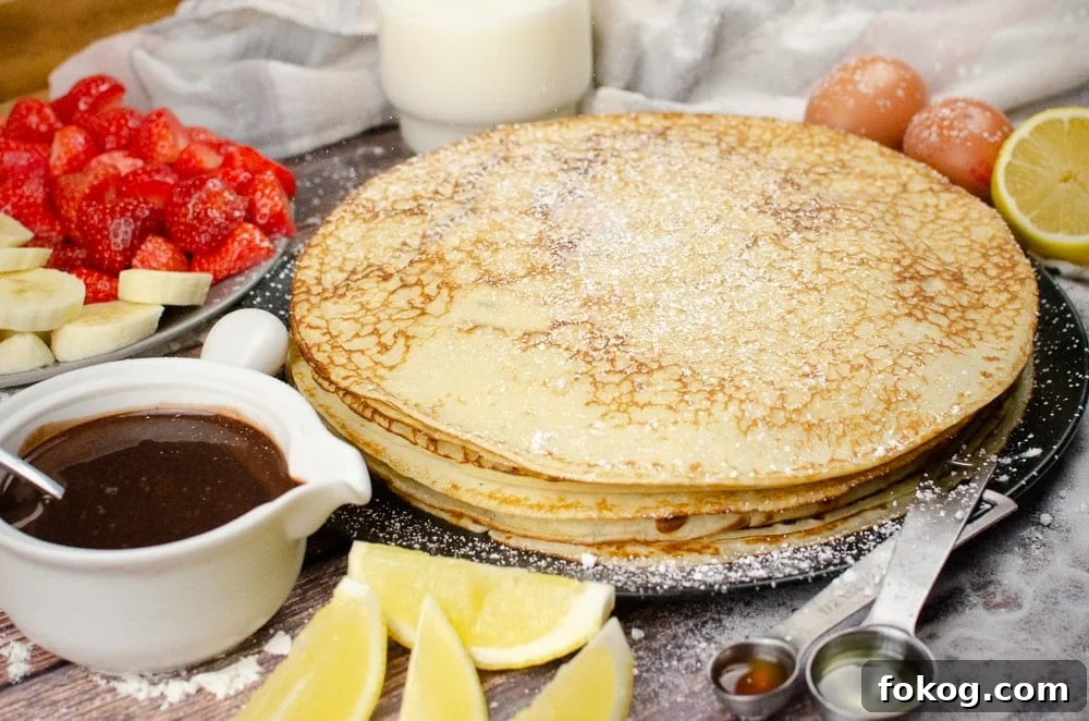 Sprinkling icing sugar over a stack of Homemade English Pancakes on a black plate on a surface of scattered sugar with a white pot of chocolate sauce, plate of chopped strawberries and bananas, a glass of milk, two eggs, slices of lemon,½ tablespoon of vegetable oil and ½ teaspoon of vanilla extract on the side
