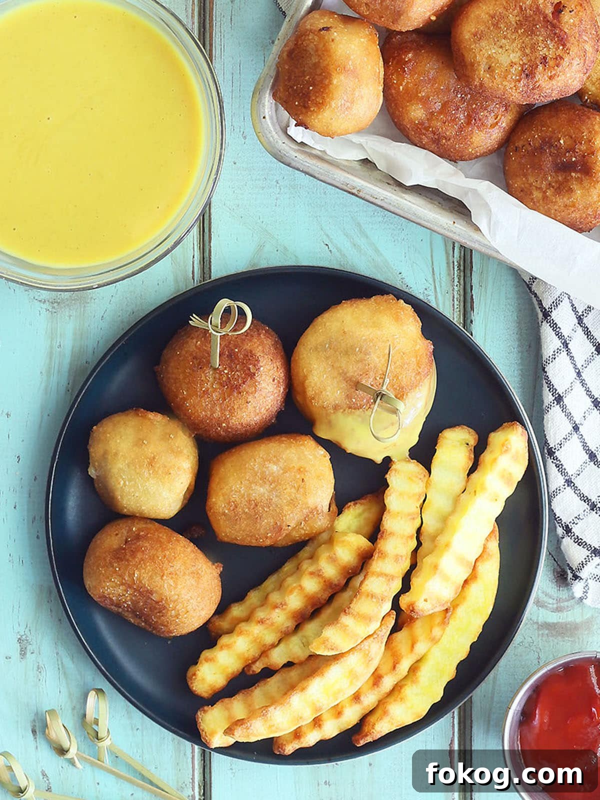 Mini Corn Dog Bites Recipe: Crispy, Bite-Sized Snack Ideas 2 Five mini corndog bites on a plate with crinkle cut French fries. Half of one corndog bite is coated in honey mustard sauce. A dish of honey mustard sauce, ketchup, toothpicks, and a platter of corndog bites are to the side of the plate.