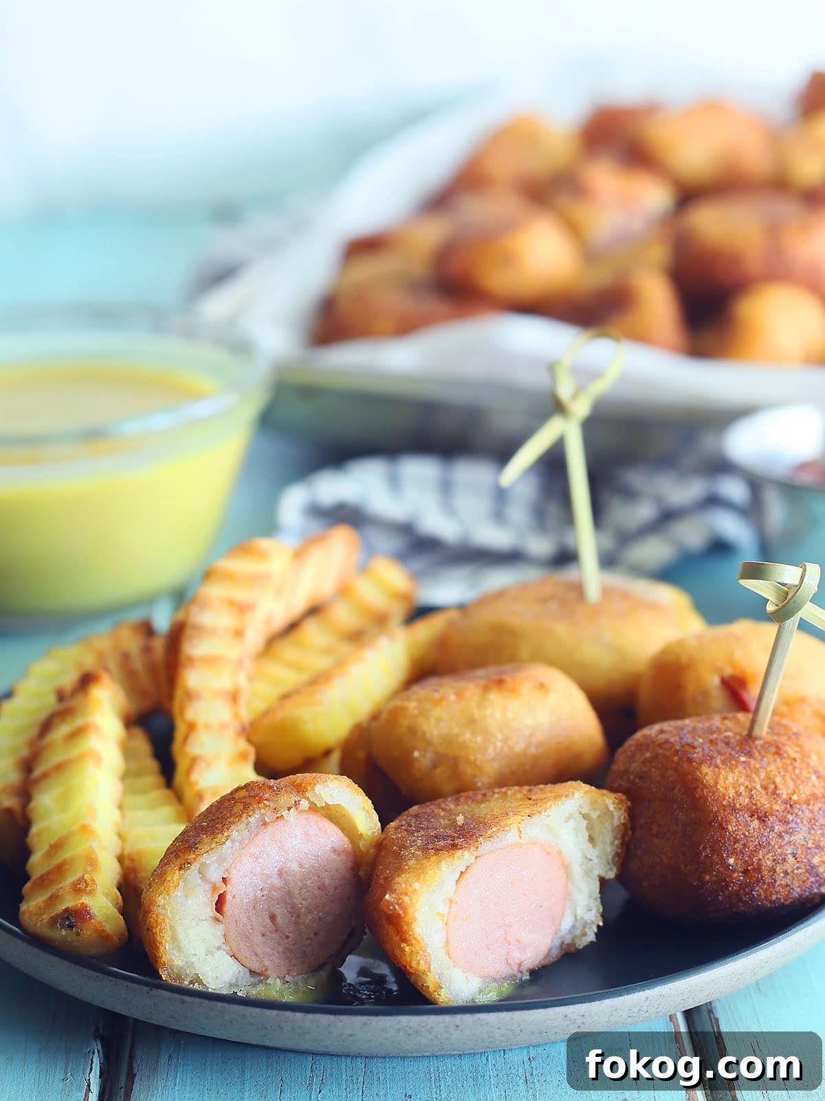 Mini Corn Dog Bites Recipe: Crispy, Bite-Sized Snack Ideas 31 Mini corndog bites on a plate with crinkle cut French fries. One corndog bite cut in half. Platter of corndog bites and a small dish of honey mustard sauce in the background.
