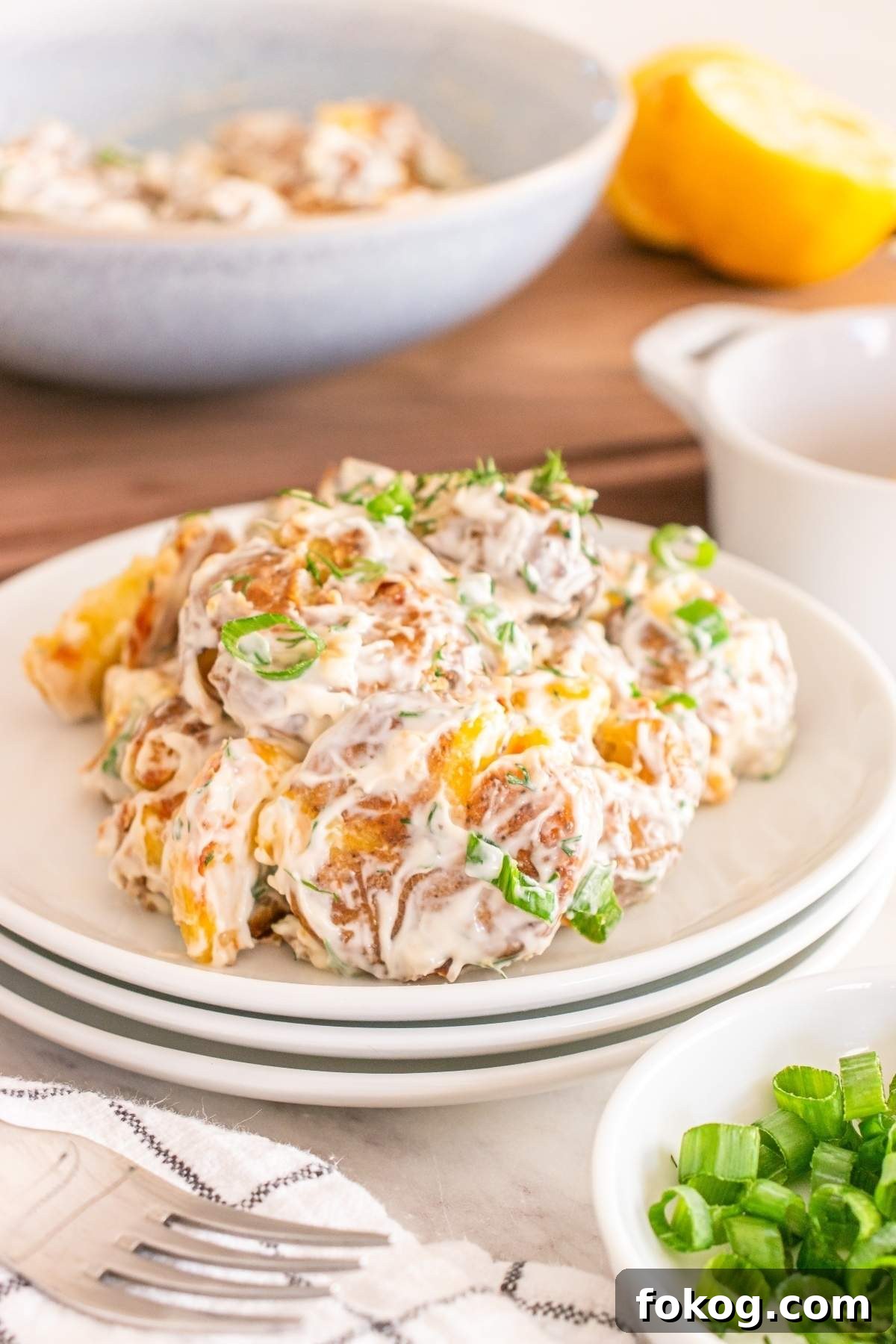A plate of creamy smashed potato salad garnished with chopped herbs. A sliced lemon and green onions are visible in the background.