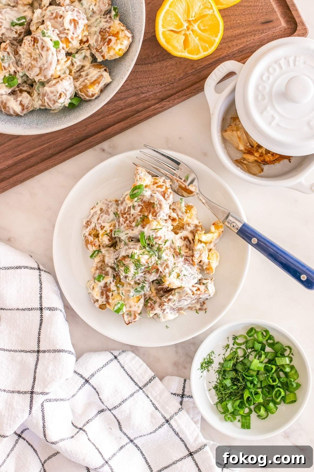 A plate of creamy smashed potato salad garnished with herbs sits next to a bowl of sliced green onions and a lemon half, all elegantly arranged on a wooden and white cloth background.