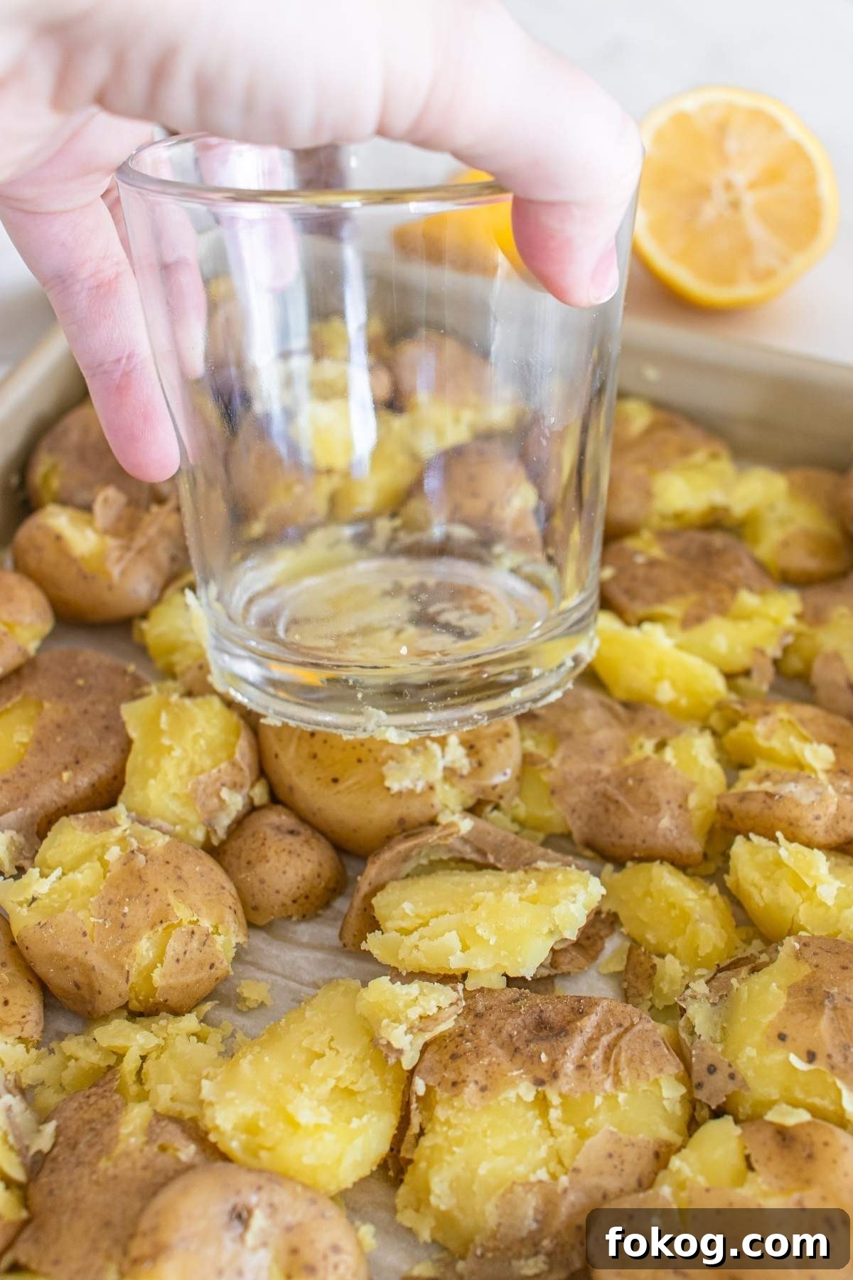 A hand uses a glass to smash roasted potatoes on a baking sheet, creating the perfect base for a delightful smashed potato salad. A halved lemon is visible in the background, hinting at the citrusy zest to come.