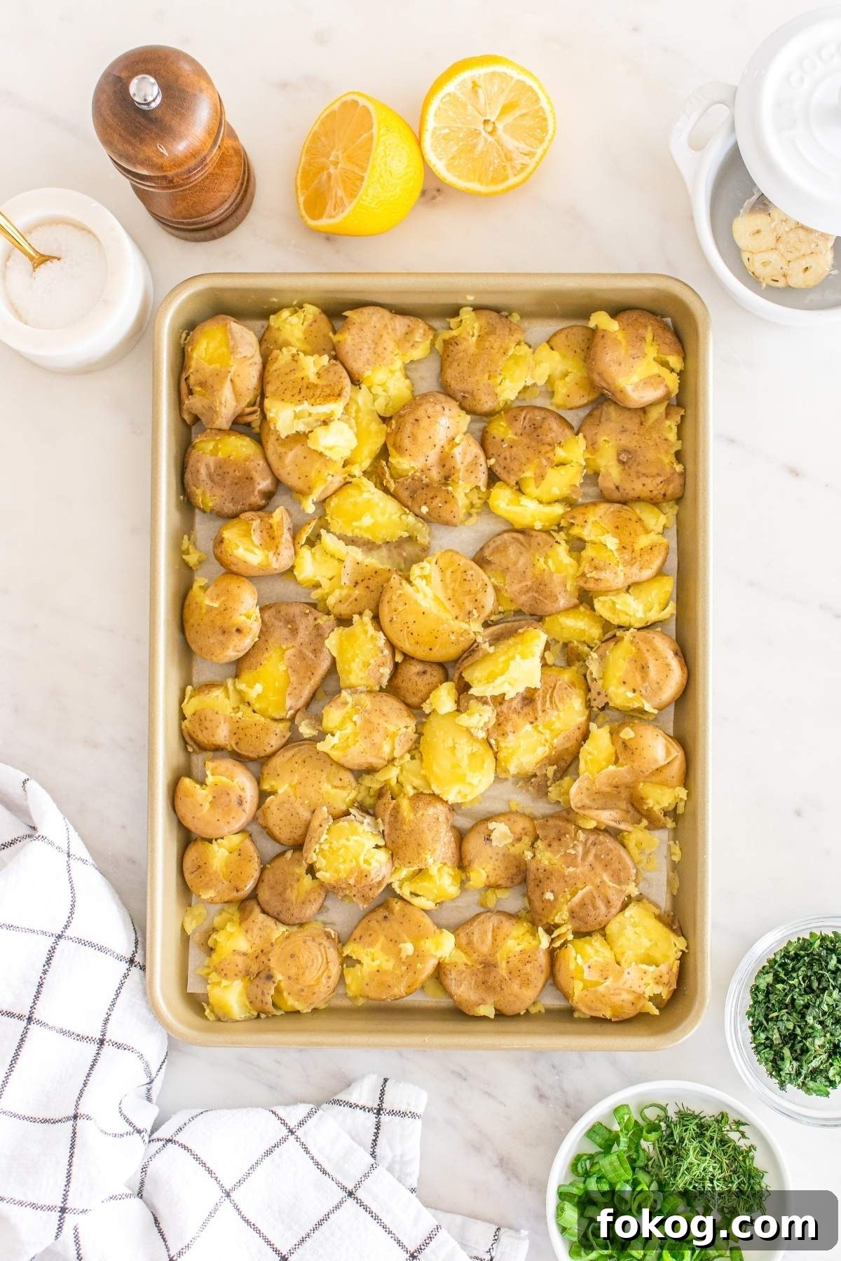 A baking tray of smashed potatoes, resembling a rustic smashed potato salad, is drizzled with seasoning and surrounded by halved lemons, a pepper grinder, and small bowls of chopped herbs on a pristine white cloth atop a marble surface.
