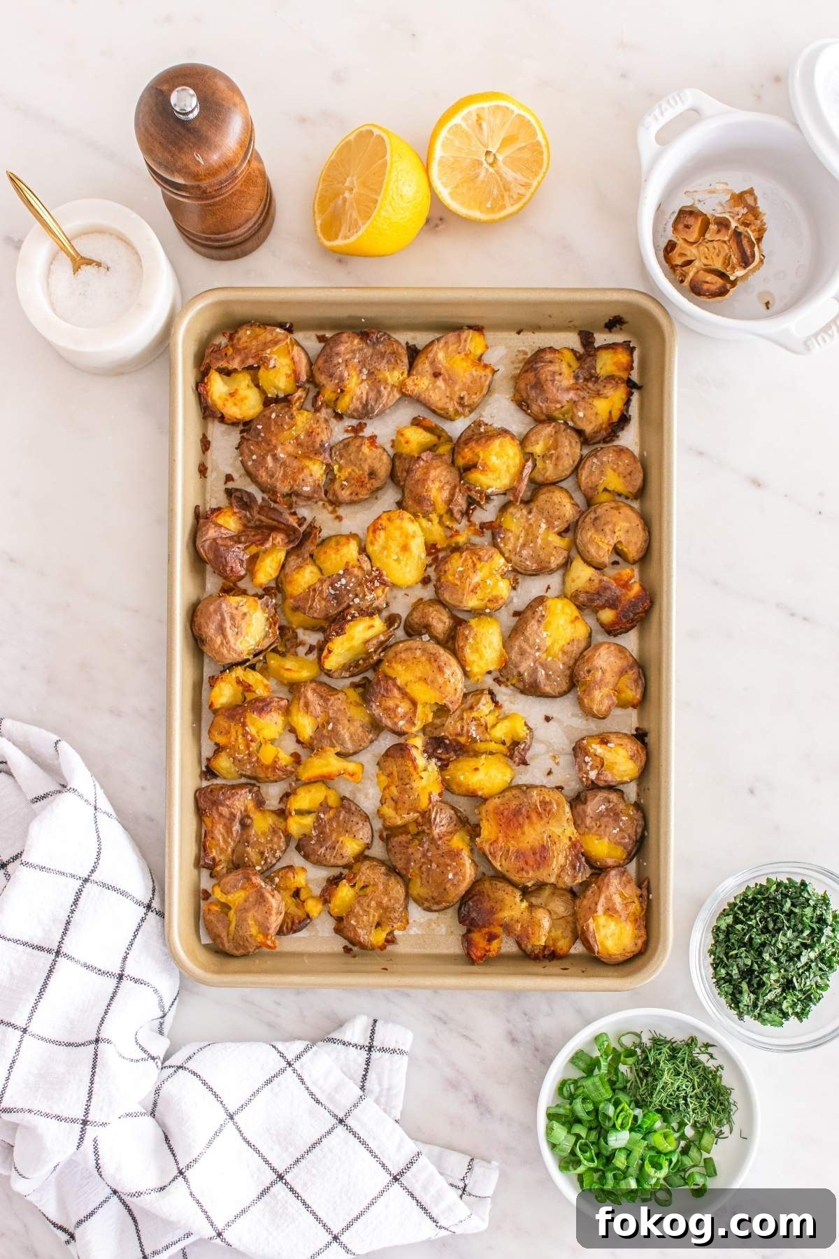 A tray of roasted potatoes, perfect for a smashed potato salad, is enveloped by a black-and-white napkin. It is accompanied by salt, pepper, halved lemon, dried garlic, and bowls of chopped herbs.