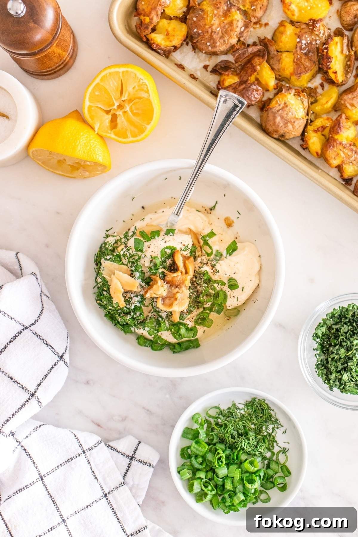 A bowl of dip garnished with green onions, herbs, and seasoning is surrounded by fresh lemon and a tempting smashed potato salad on a tray, along with various condiments on a marble surface.