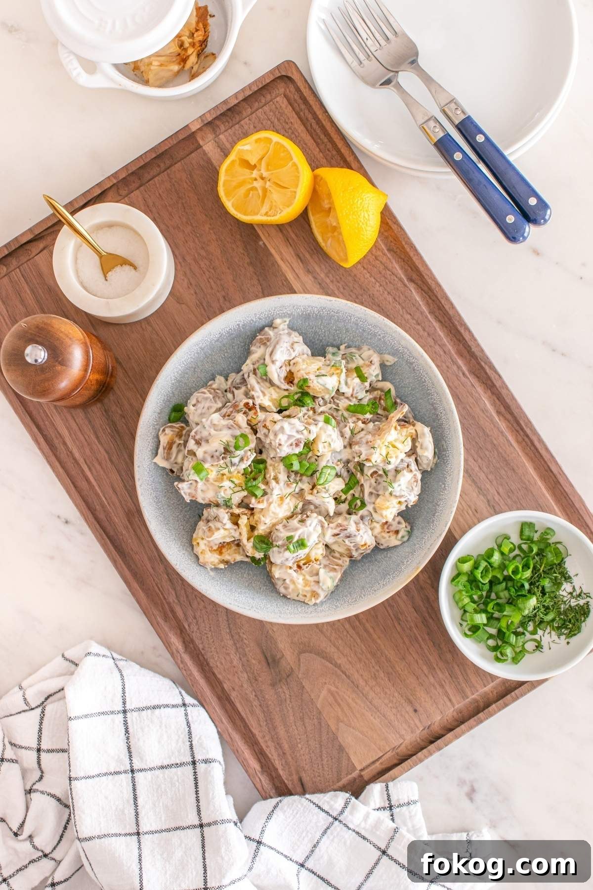 A bowl of smashed potato salad with green onions sits on a wooden board. Nearby are lemon halves, a small bowl of chopped green onions, salt, pepper, and forks on plates. A checkered napkin is visible beside the delightful display.