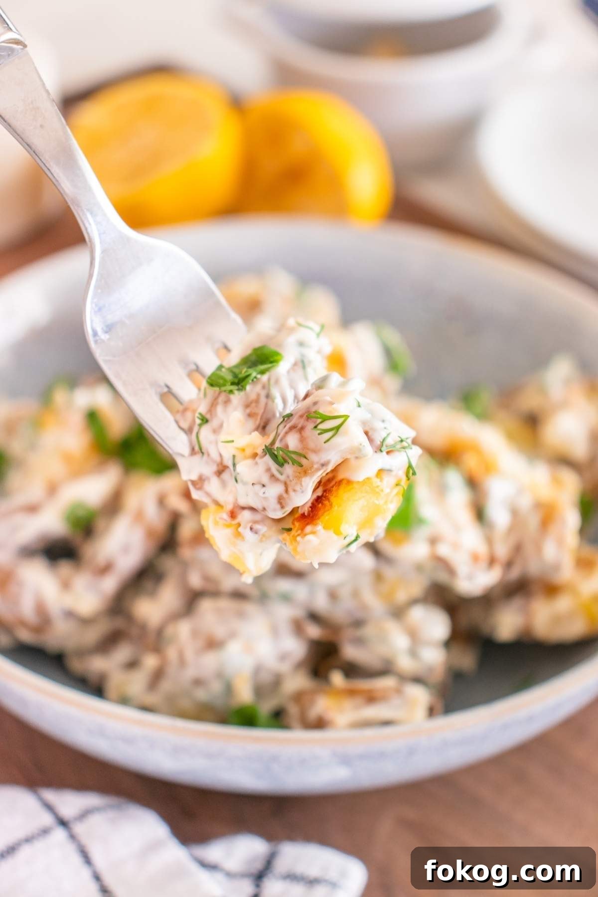 A fork holds a bite of creamy smashed potato salad over a bowl brimming with the same dish. Lemons and a napkin gracefully adorn the background.