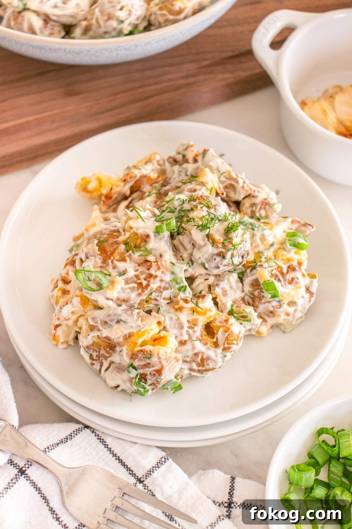 A plate of smashed potato salad, garnished with fresh herbs and green onions, sits invitingly on a checkered napkin with a fork beside it.