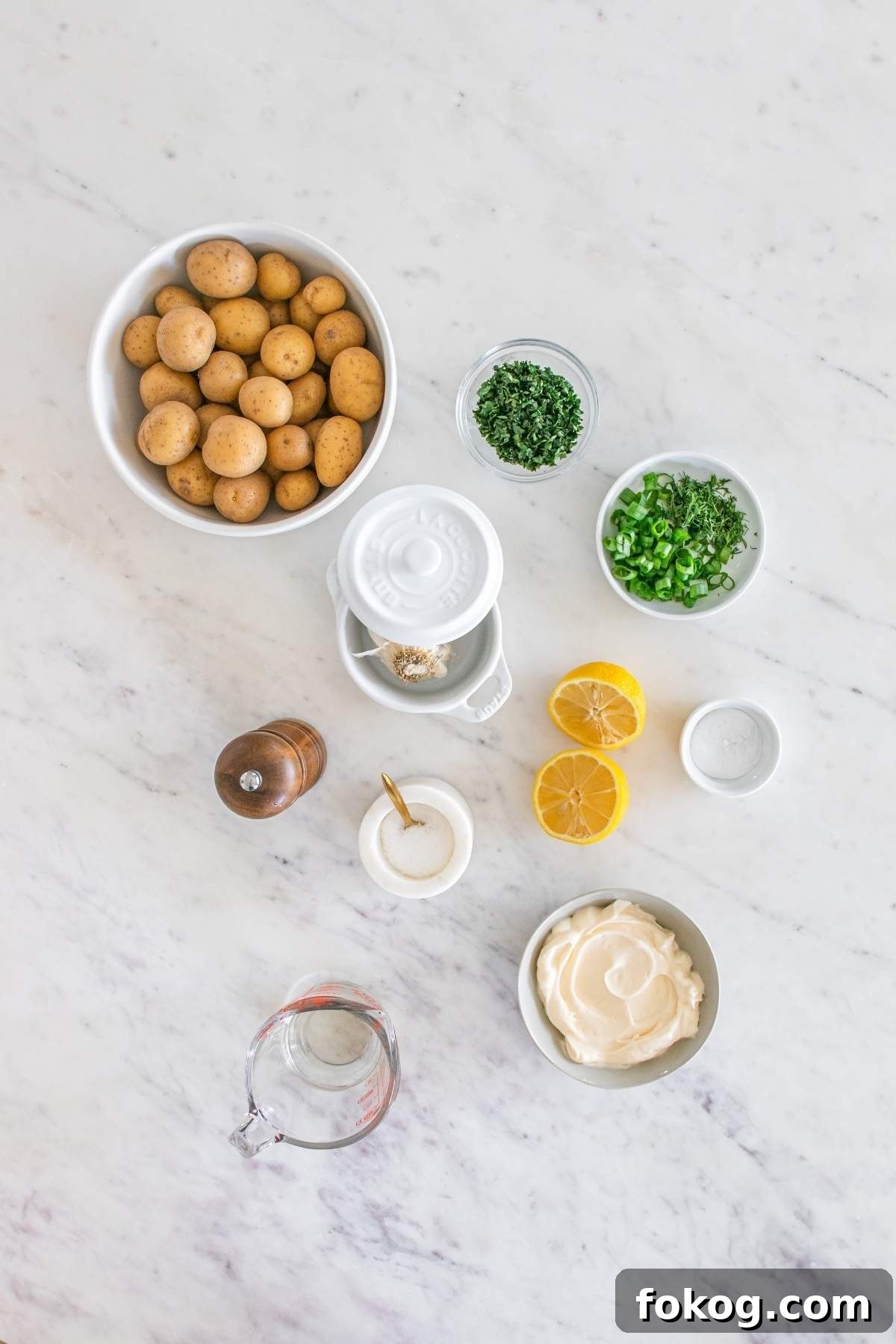 Top view of ingredients for a smashed potato salad on a marble countertop, featuring a bowl of small potatoes, chopped herbs, lemon halves, a bowl of mayonnaise, a pepper grinder, and various measuring tools.