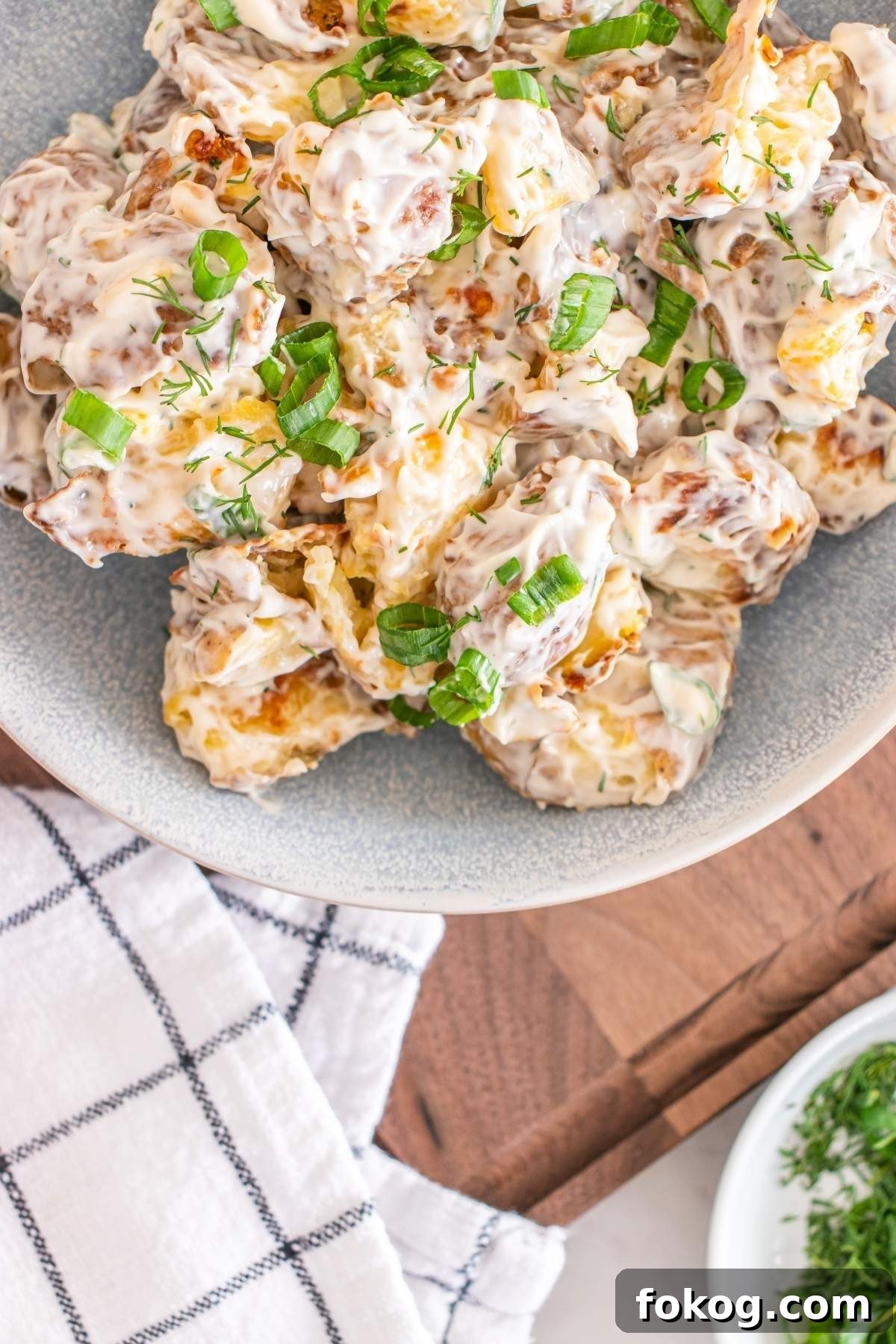 A bowl of creamy smashed potato salad with green onions on top sits on a wooden surface next to a checkered napkin and a small dish of herbs.