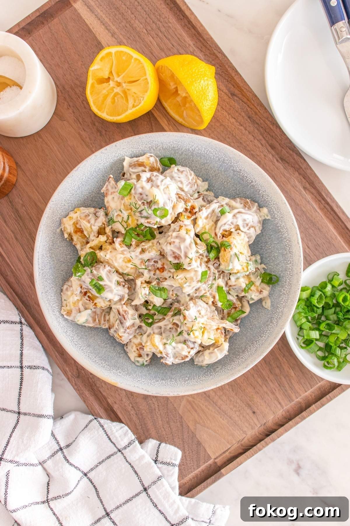 A bowl of creamy smashed potato salad garnished with green onions sits on a wooden board, surrounded by halved lemons and a bowl of extra chopped green onions.