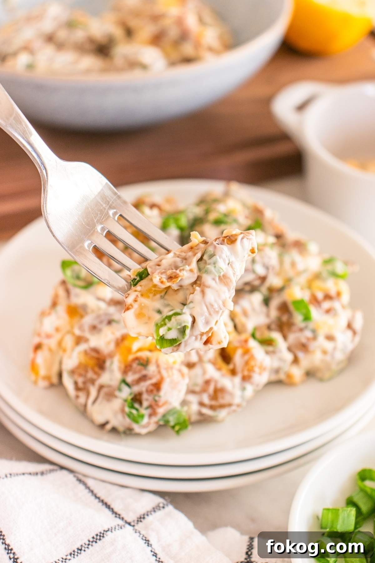 A fork holds up a forkful of creamy smashed potato salad with chopped green onions, with more salad on a white plate and ingredients in the background.