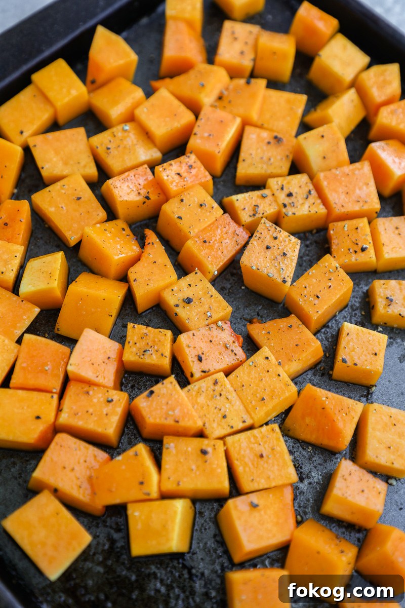 Diced butternut squash tossed in olive oil, salt and black pepper sitting are a dark baking sheet pan