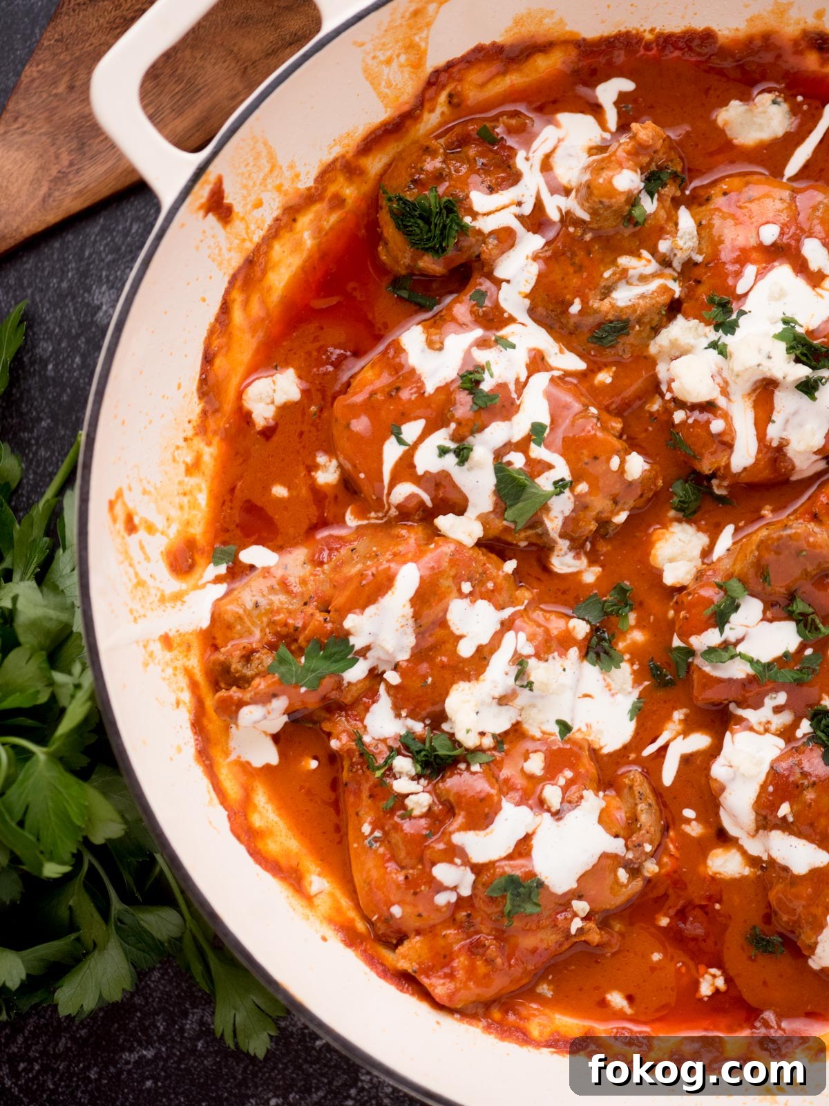 Crispy Buffalo Chicken Thighs Recipe for Dinner 2 buffalo chicken thighs in a skillet surrounded by fresh parsley, a serving utensil, and a kitchen towel
