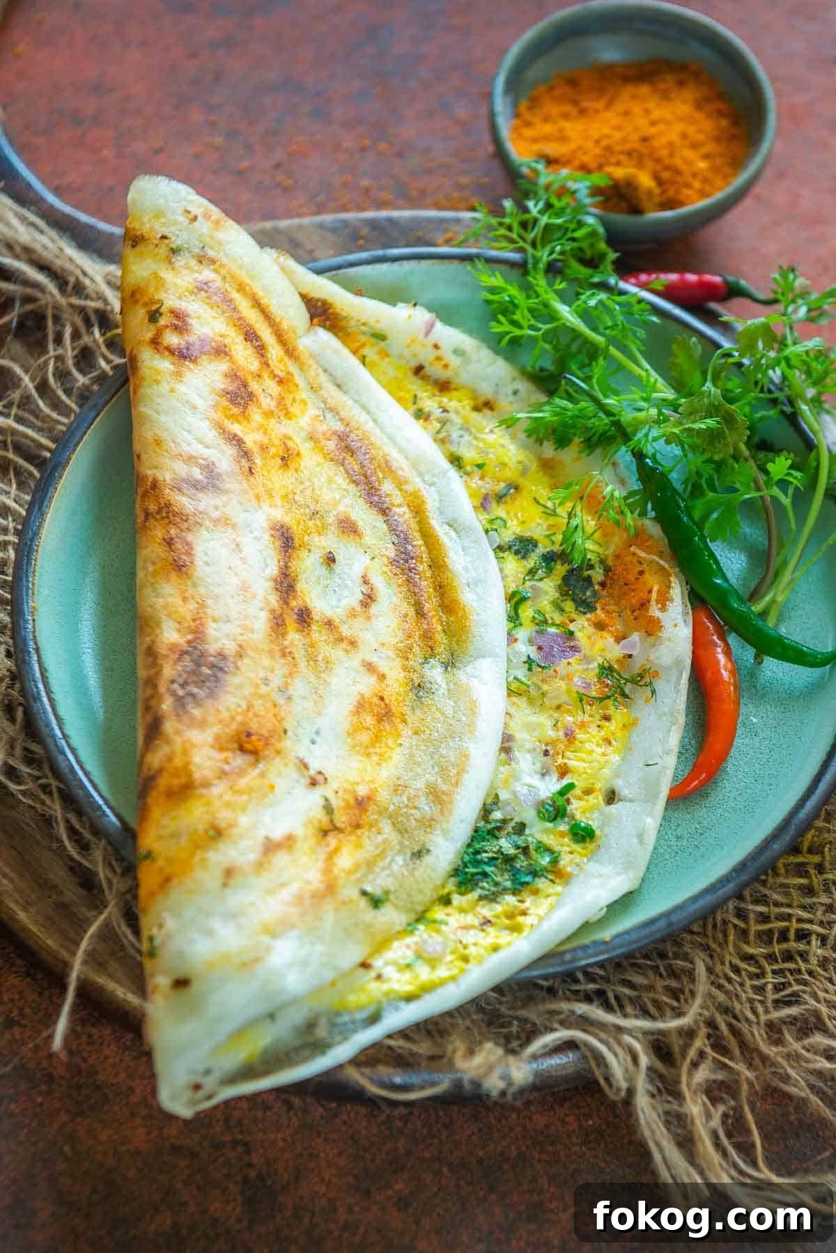 Crispy Egg Dosa (Mutta Dosa) Recipe for Perfect Breakfast 2 A crispy Egg Dosa served on a green plate with red chilies, cilantro, and a small bowl of podi on the side.