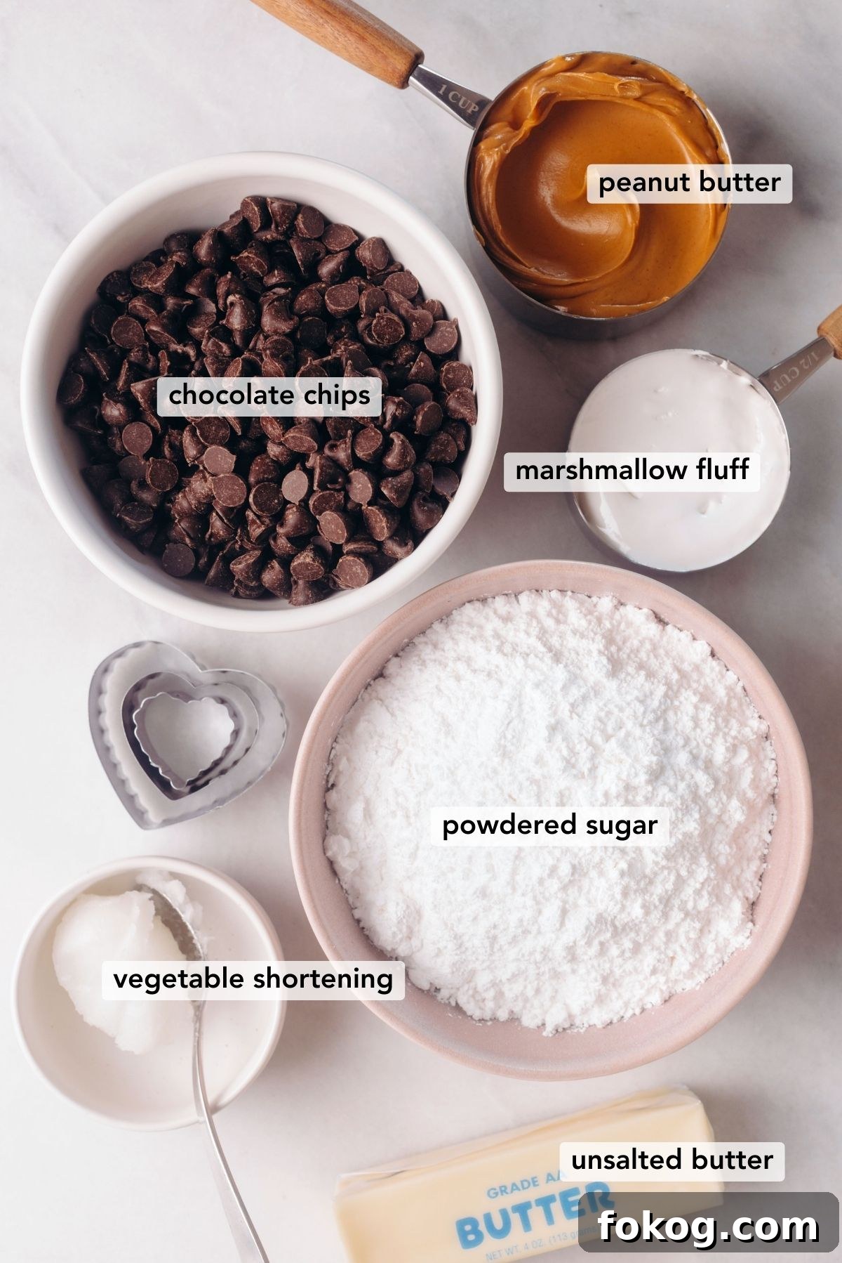 Ingredients needed for chocolate covered peanut butter eggs with three heart-shaped cookie cutters on a white marbled background.
