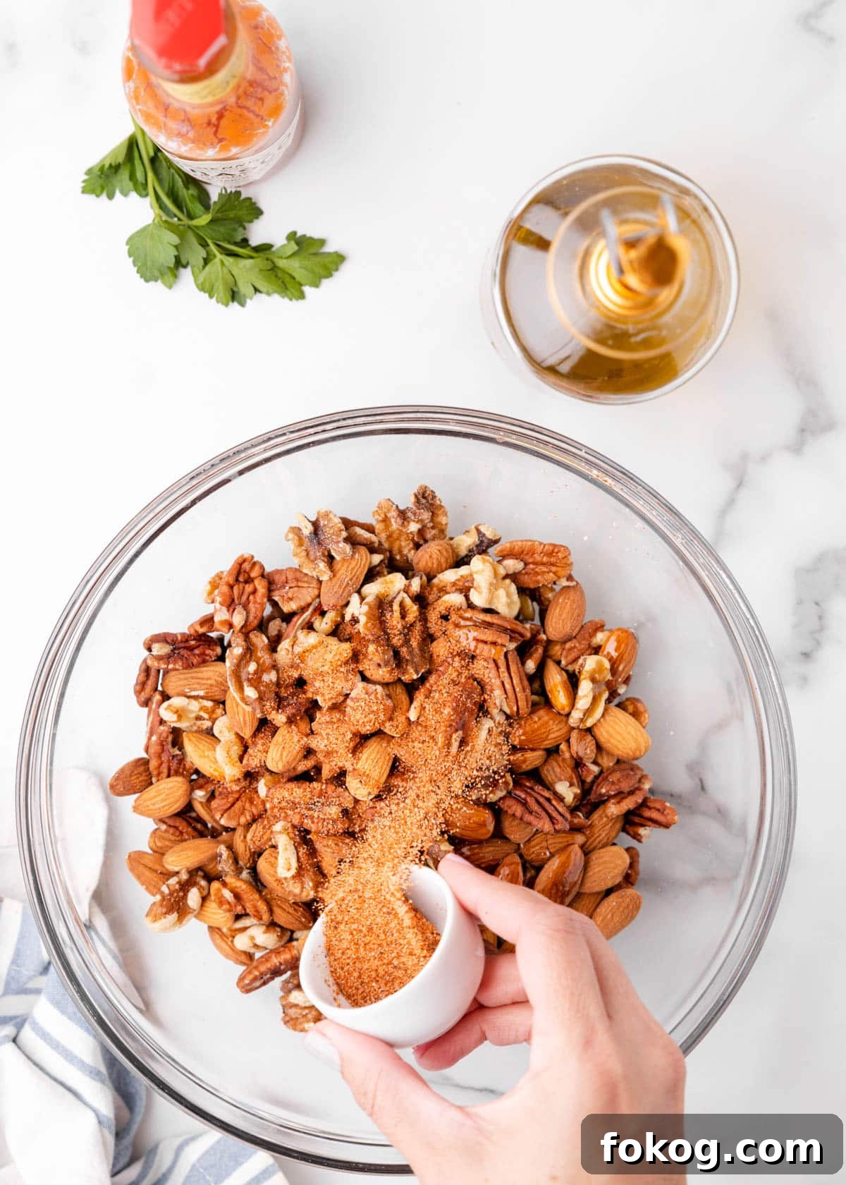 overhead photo showing cajun seasoning being poured into a glass bowl of mixed nuts on a marble board