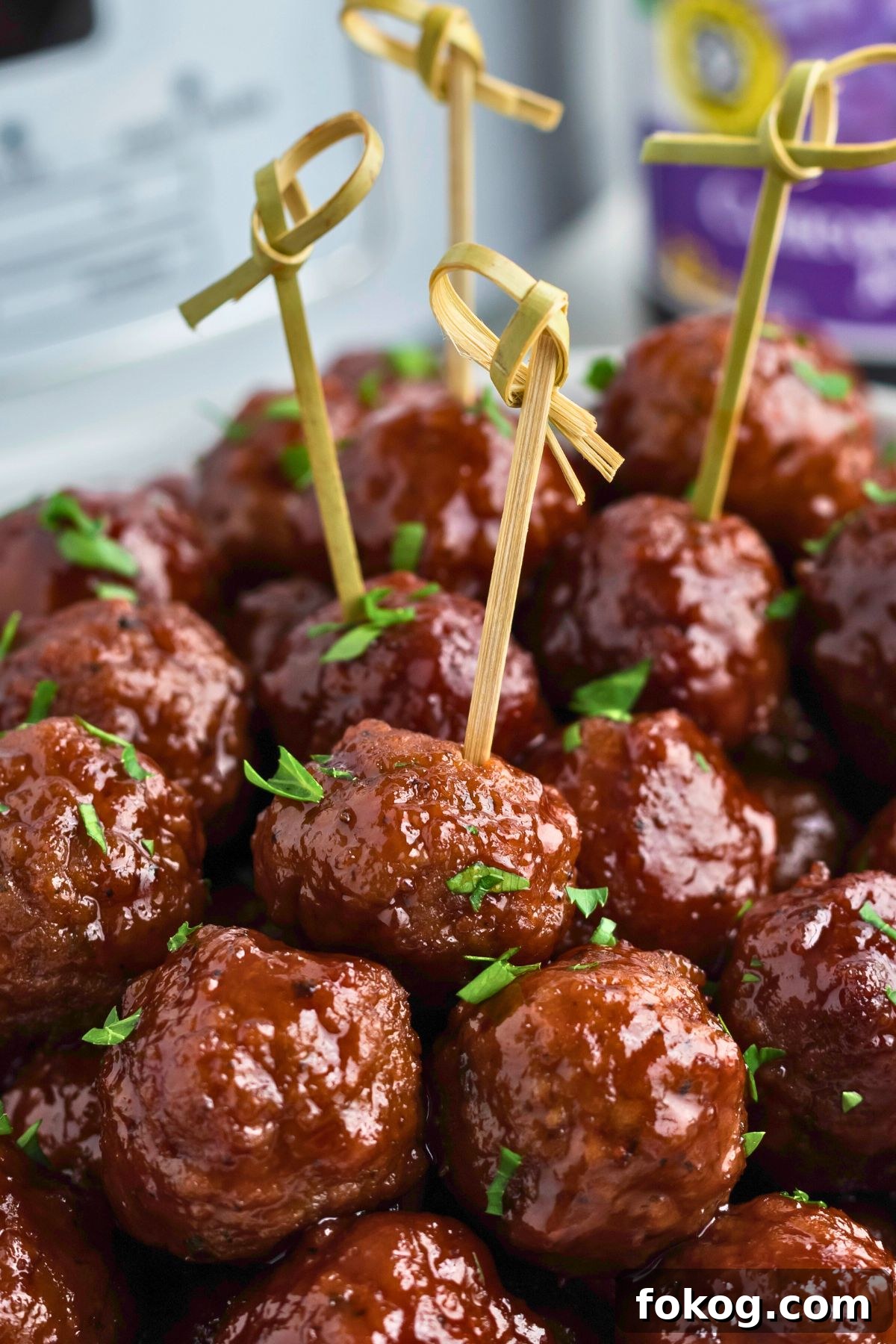 Sweet Grape Jelly Meatballs with Tangy Glaze Recipe 2 Grape jelly meatballs stacked with some skewered, in front of a slow cooker and jar of grape jelly.