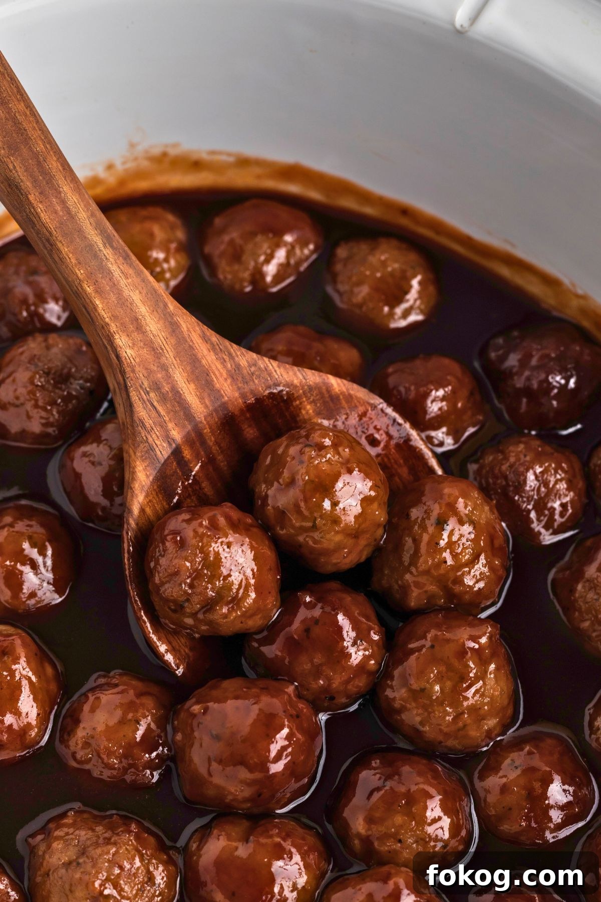 Sweet Grape Jelly Meatballs with Tangy Glaze Recipe 7 Grape jelly meatballs in a white crock pot being scooped up to serve with a wooden spoon.