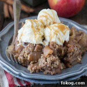 Slow-Cooker Apple Crisp with Cinnamon Oat Topping 7 A plate of crock pot apple crisp topped with three scoops of vanilla ice cream and drizzled with caramel sauce, with a fork on the side. An apple and cinnamon sticks are in the background.
