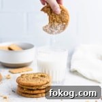 A stack of graham cracker cookies next to a glass of milk. A hand from the top of the frame is holding a cookie over the glass as milk drips off it.