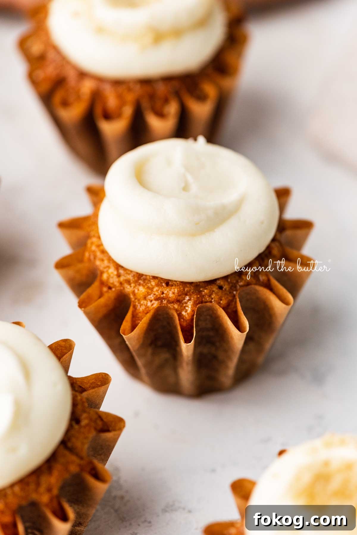 Carrot cake cupcakes topped with cream cheese frosting on a white background | © Beyond the Butter®