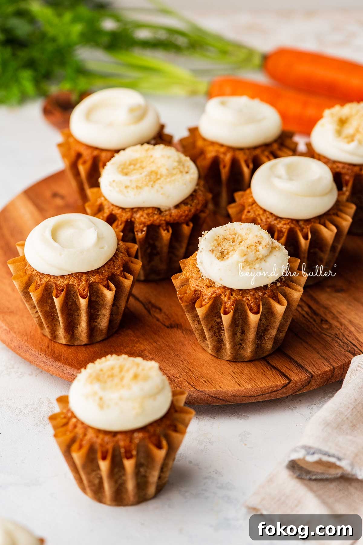 Super moist carrot cake cupcakes with cream cheese frosting placed on a wood cake stand on a white background with some carrots | © Beyond the Butter®