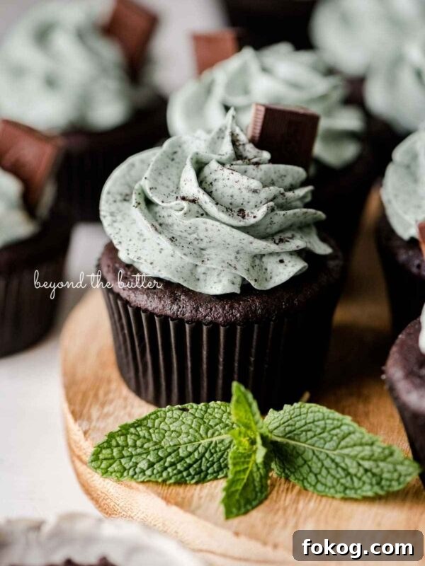 Mint chocolate cupcakes topped with mint chocolate chip frosting and an Andes mint on a wood circular tray with mint leaves as decoration.