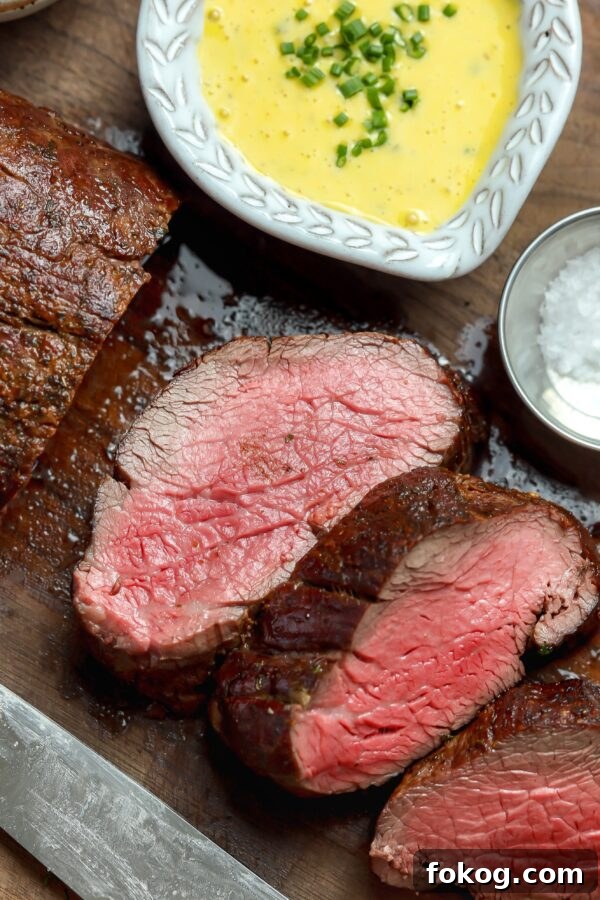 Perfectly seared and thinly sliced pieces of slow roasted tenderloin glistening on top of a dark wood cutting board.