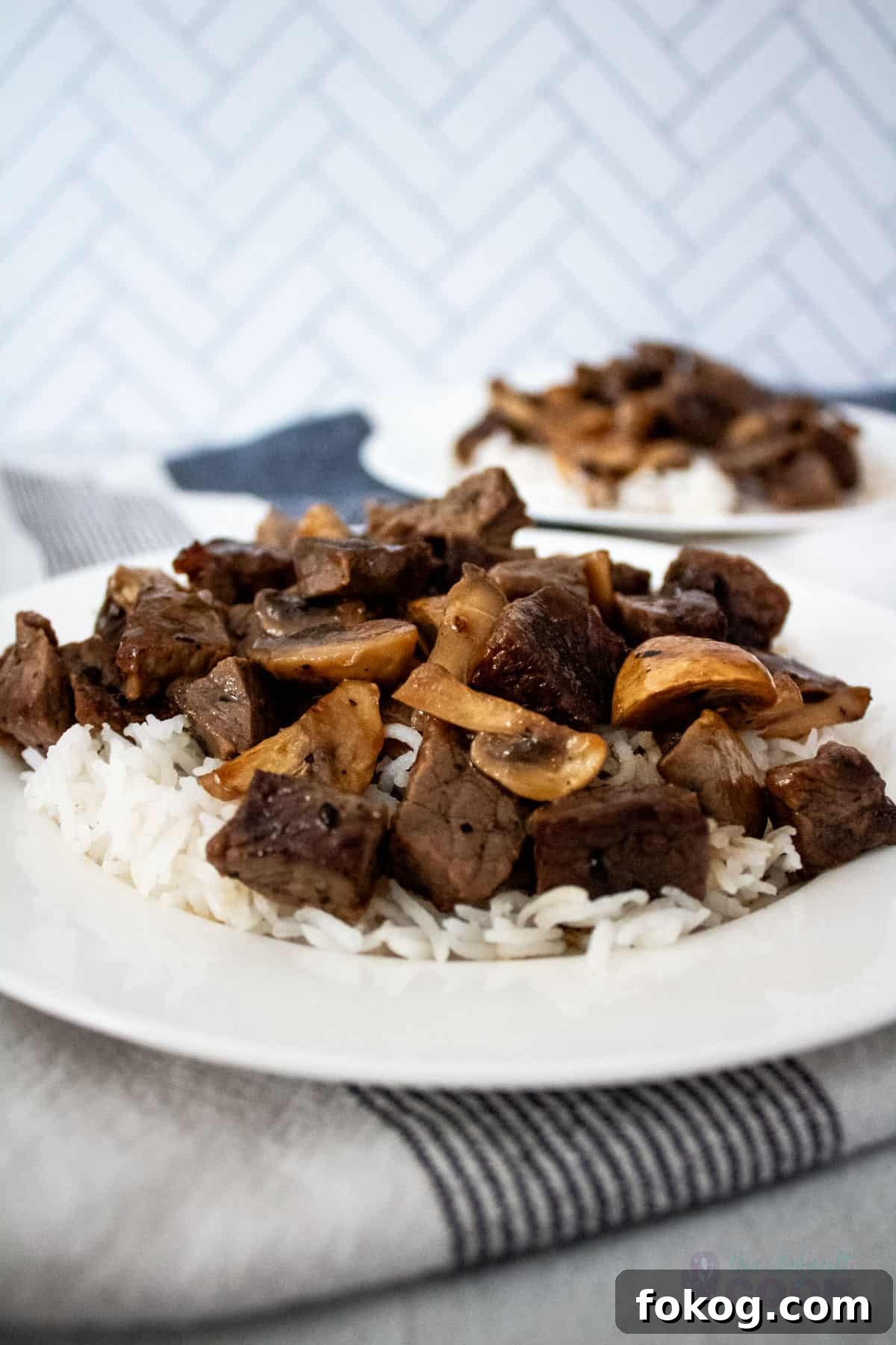 Two plates of rice topped with steak bites and mushrooms on a blue and white towel with chevron tile in the background.
