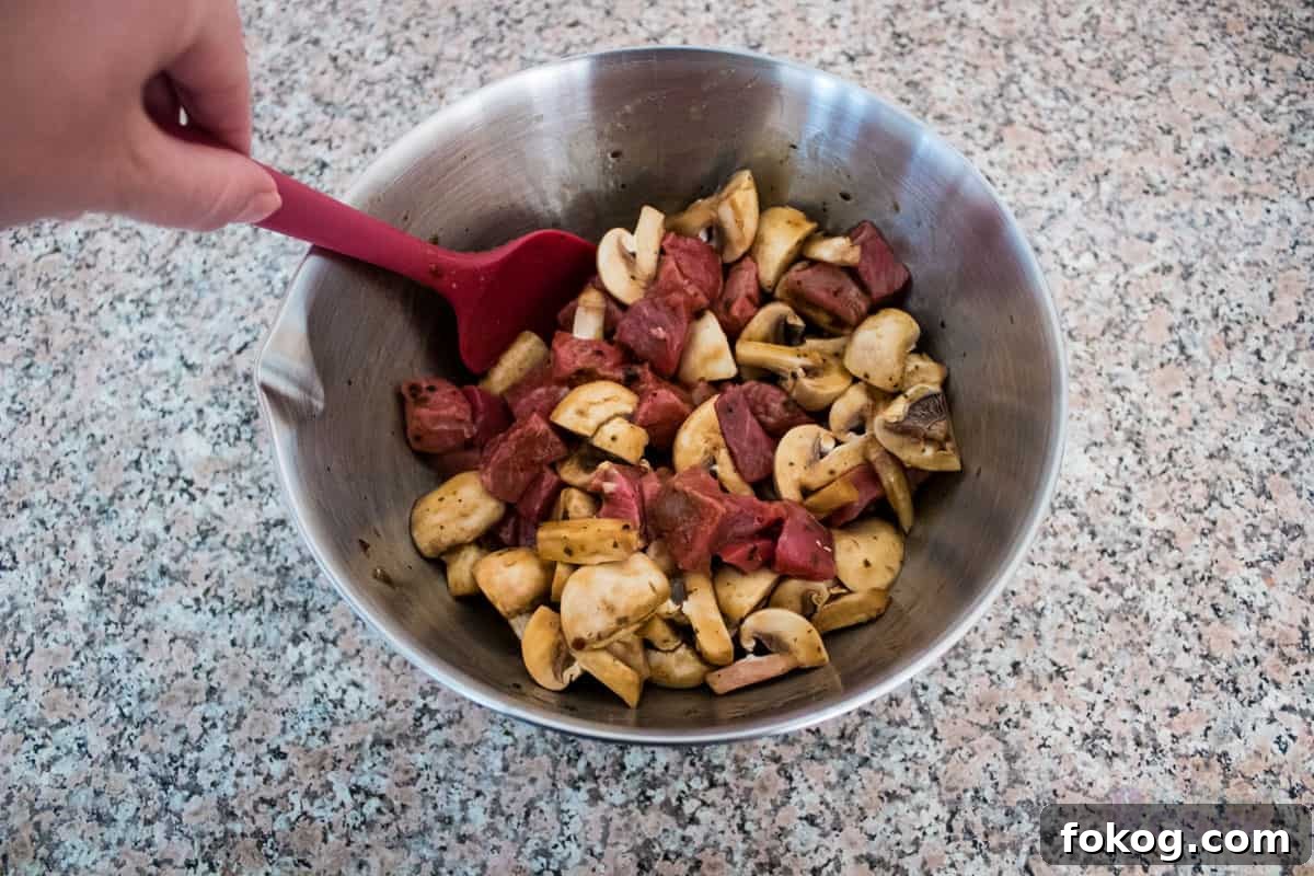 A steel bowl with mushrooms and steak coated in a sauce, being stirred by a hand holding a red spatula.