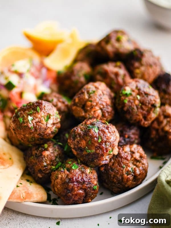 Close-up side view of browned Greek meatballs garnished with chopped parsley on a light beige plate with pita bread and lemon wedges.
