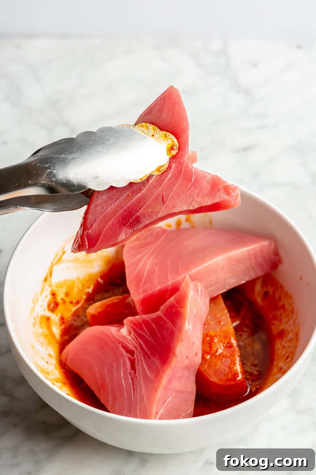 Chili-Lime Seared Ahi Tuna with Cilantro-Lime Sauce 5 A pair of tongs holding up a tuna steak over a white bowl with chili-lime marinade.
