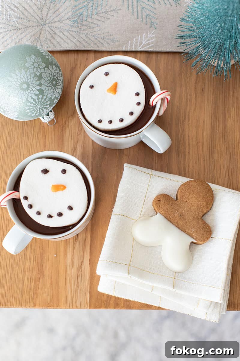 Table top with hot chocolate and snowmen marshmallows, a gingerbread cookie on a white and gold napkin and a sage ornament 