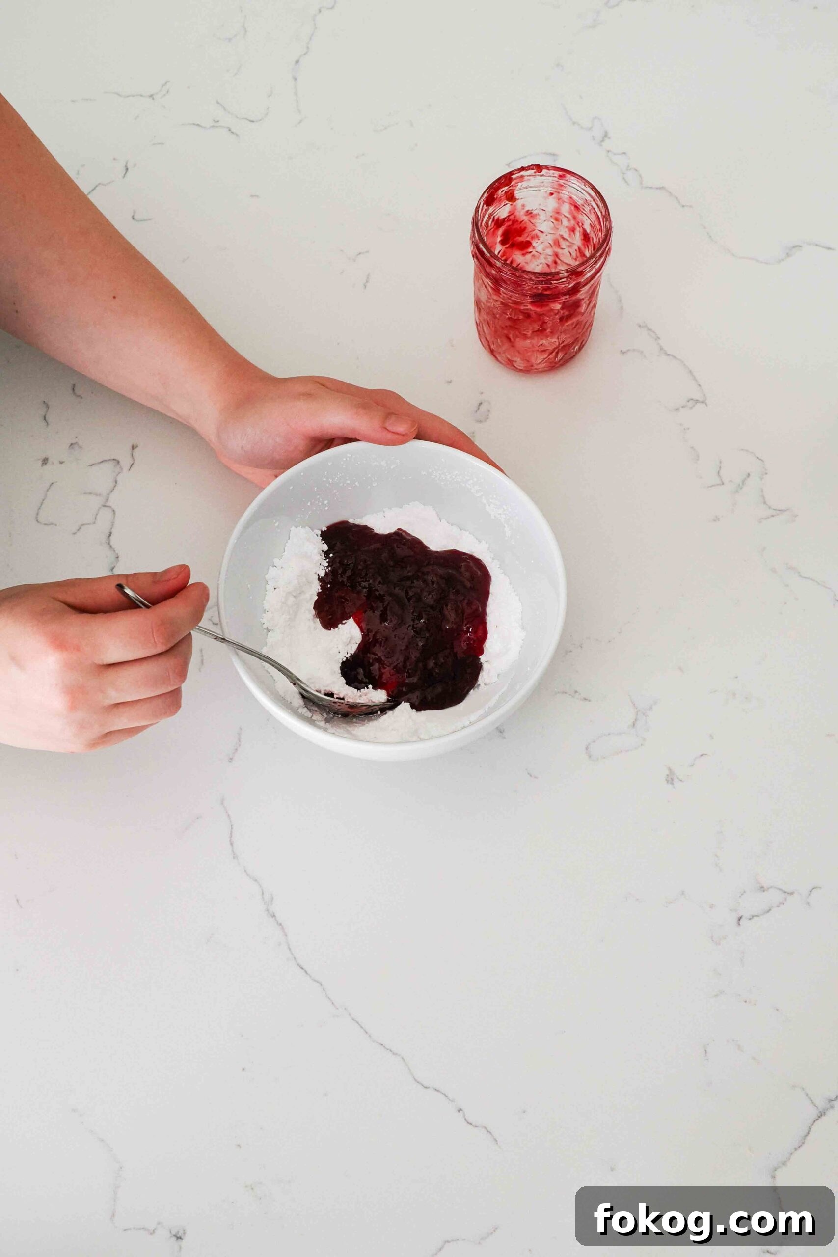 Two hands begin stirring together cherry jam and powdered sugar in a white bowl.
