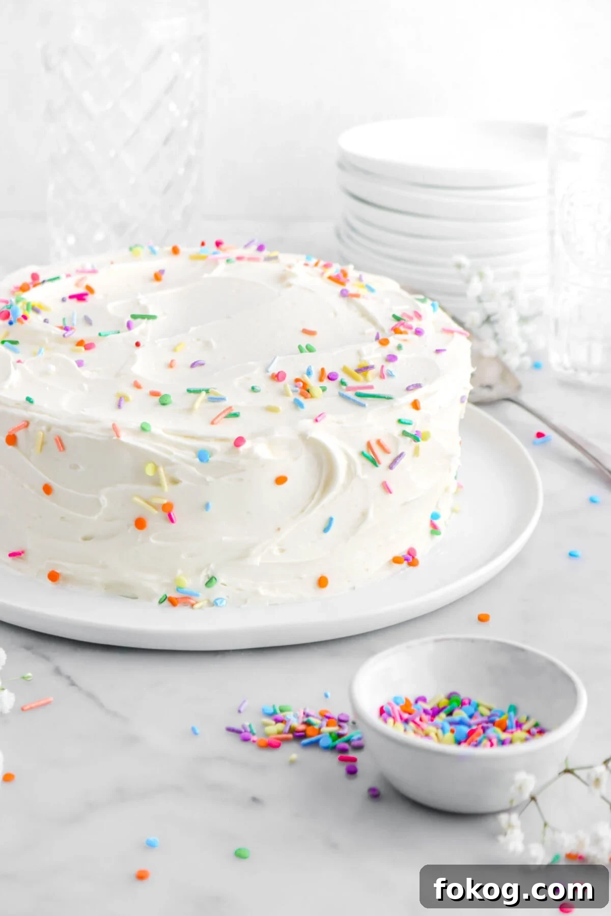 Classic Vanilla Cake with Smooth Buttercream Frosting 3 cropped close up of frosted cake with sprinkles on top and sides of cake, with bowl of sprinkles and white flowers around on marble surface.