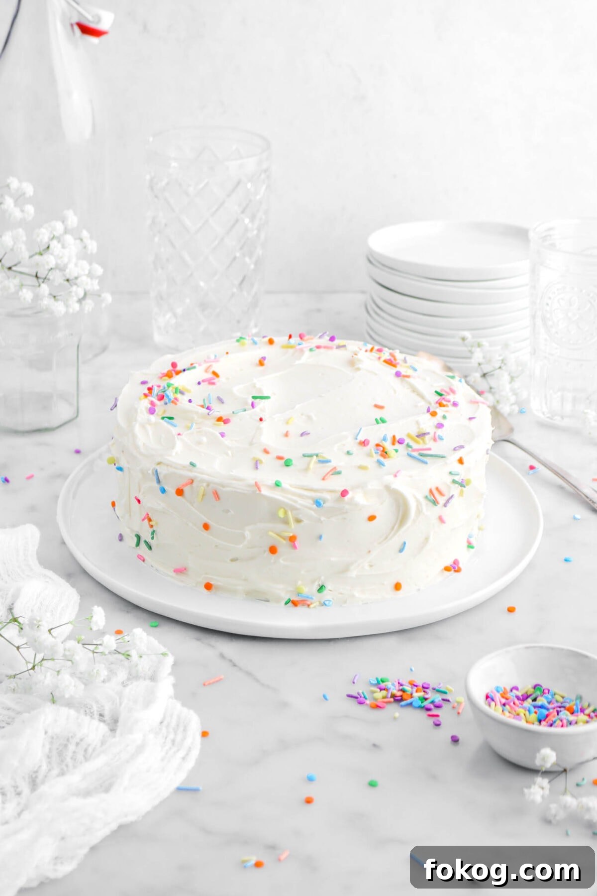 Classic Vanilla Cake with Smooth Buttercream Frosting 18 front shot of frosted cake with sprinkles scattered around on marble surface, with white cheesecloth beside, white flowers, empty glasses, and stack of plates behind on marble surface.