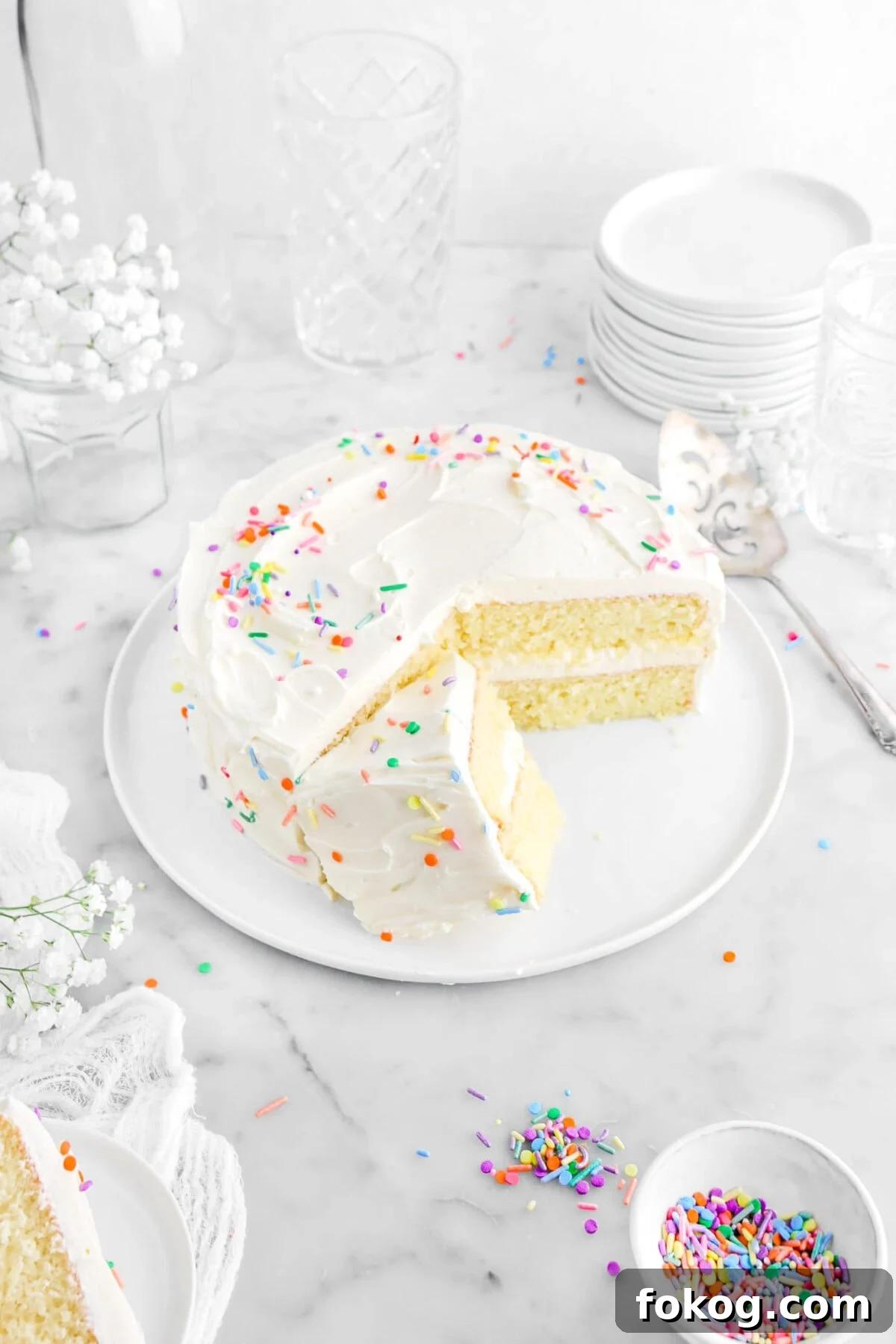 Classic Vanilla Cake with Smooth Buttercream Frosting 20 angled shot of cake with slice leaning against cake with cake knife beside, sprinkles around, white flowers, and white cheese cloth around with stack of plates and glasses behind.