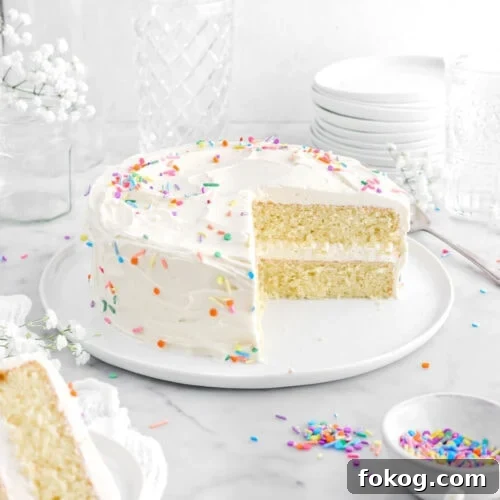 Classic Vanilla Cake with Smooth Buttercream Frosting 22 frosted vanilla cake on white plate with slice in front ons mall white plate on top of a white cheesecloth with sprinkles and white flowers around.