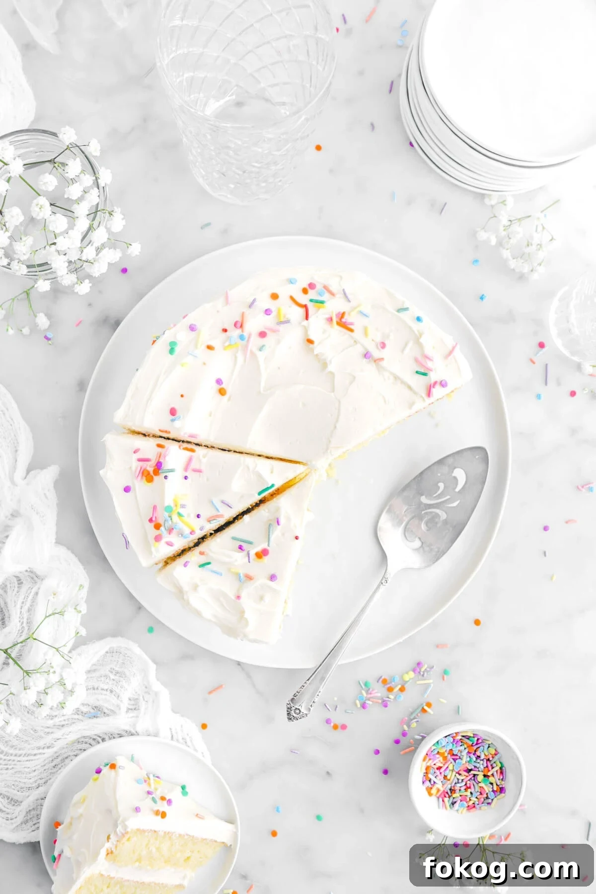 Classic Vanilla Cake with Smooth Buttercream Frosting 5 overhead shot of vanilla cake on white plate with two slices cut into and cake knife beside cake, with sprinkles, white flowers, stack of plates, and slice of cake on small white plate on top of white cheesecloth.