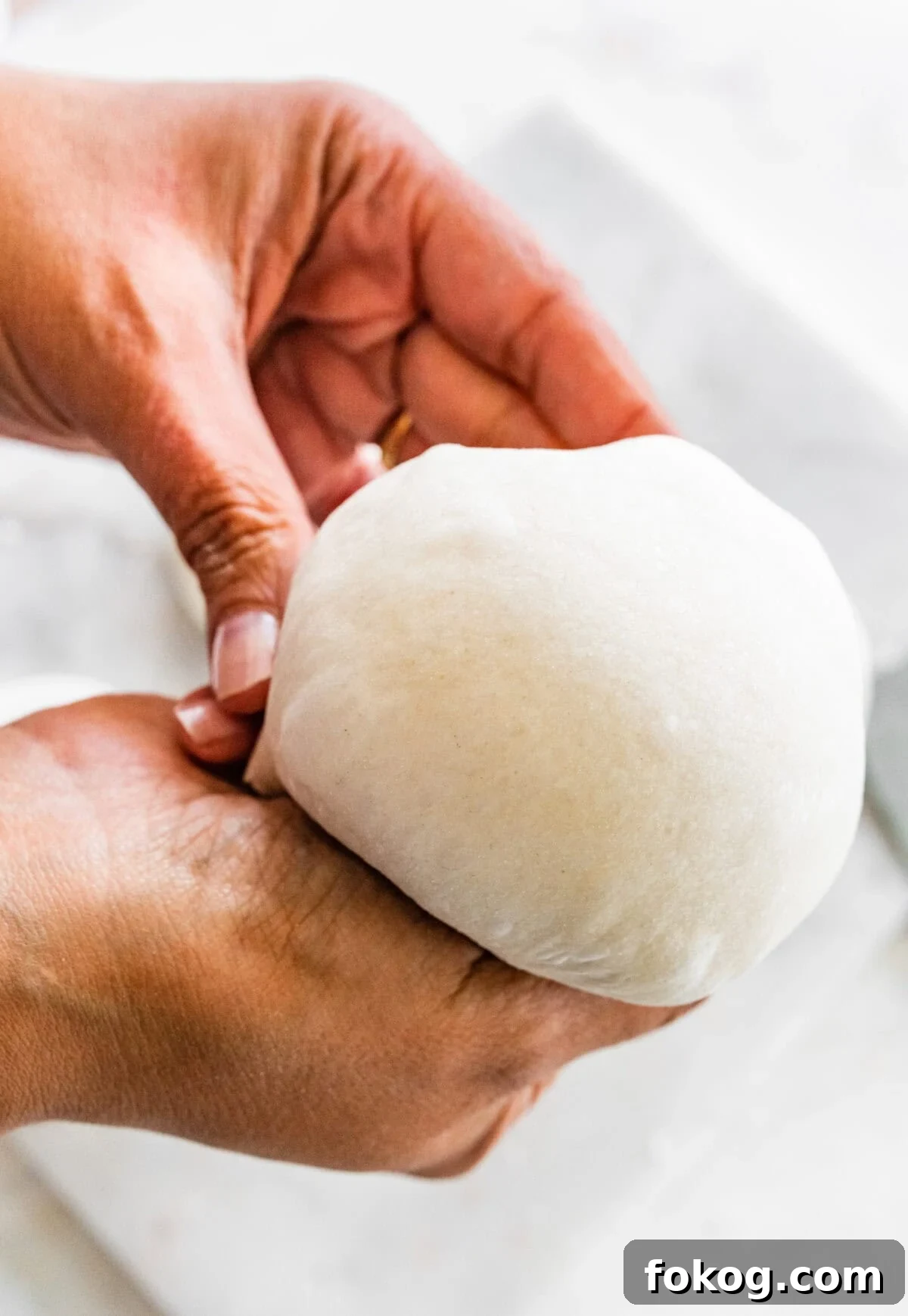 A person holding a dough ball on a counter using tuck and fold method.
