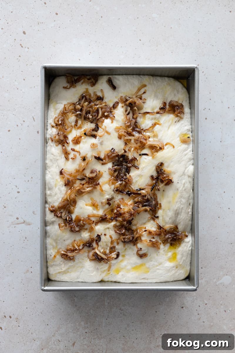 Focaccia dough in a baking pan with olive oil and caramelized shallots on top. 
